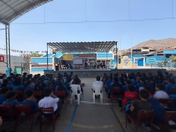 Estudiantes sentados en un patio escolar escuchan a ponentes ubicados sobre un escenario techado
