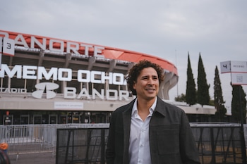Un hombre con camisa blanca y chaqueta gris posa delante de un estadio deportivo que tiene un letrero con las palabras Memo Ochoa