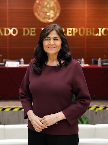 Una mujer con cabello oscuro y blusa color burdeos sonríe ligeramente, con las manos entrelazadas al frente. Detrás hay una pared de madera con letras doradas y un emblema
