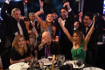 Hombres y mujeres con trajes formales levantan los brazos y sonríen sentados y de pie junto a una mesa con copas
