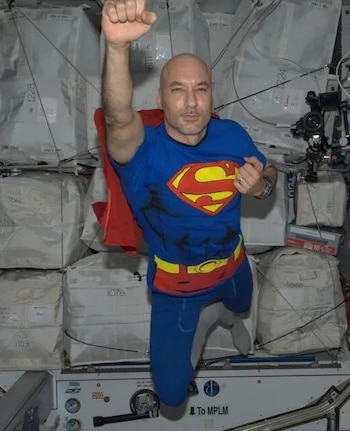 Un hombre con camiseta de Superman y capa roja flota con el puño en alto frente a paquetes blancos de carga en una nave espacial