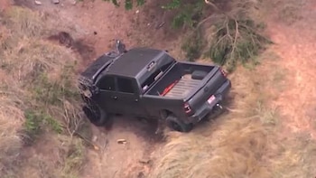 Vista aérea de una camioneta pickup oscura atascada en una zanja de tierra con vegetación seca y el frente del vehículo visiblemente dañado