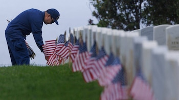 Un hombre uniformado se inclina para colocar pequeñas banderas de Estados Unidos frente a una fila de lápidas blancas en un campo verde