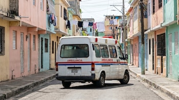 Ambulancia blanca estacionada en una calle estrecha de República Dominicana, flanqueada por edificios coloridos y ropa tendida en el fondo.