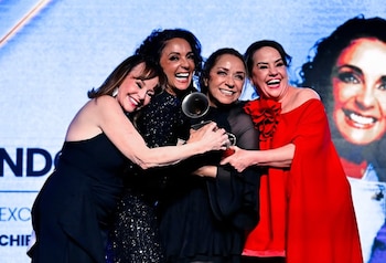 Cuatro mujeres en vestidos de gala se abrazan en un escenario. Una sostiene un trofeo. Al fondo, una pantalla muestra texto y el rostro de una mujer