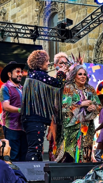 Cuatro personas en un escenario. Una mujer de cabello gris recibe una corona puntiaguda. La acompaña otra persona colocando la corona. Un hombre con sombrero y gafas sonríe