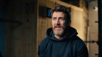 Aldo Kane, hombre con barba y cabello castaño, usa una sudadera azul oscura. Mira hacia arriba con expresión serena. Fondo borroso con paredes de madera y barras