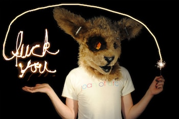 Persona con máscara peluda de animal y ojos naranjas brillantes, vistiendo camiseta clara, sujeta una bengala que traza "fuck you" con luz