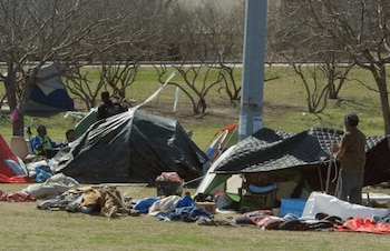 Vista de un campamento con varias tiendas de campaña y objetos personales esparcidos en un césped verde, con árboles desnudos al fondo y personas cerca de las tiendas