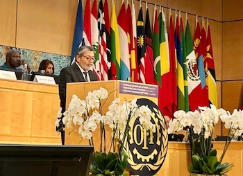 Gerardo Martínez habla desde un atril ante un fondo de banderas internacionales. Se ven arreglos de orquídeas blancas y el logotipo de la OIT en el escenario