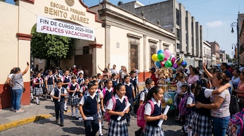Numerosos niños de primaria en uniforme salen de una escuela en México. Una pancarta dice "FIN DE CURSO 2025-2026 ¡FELICES VACACIONES!". Padres esperan a los alumnos.