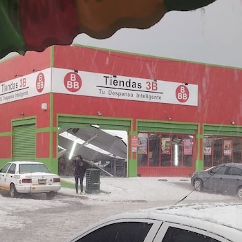 Una tienda Tiendas 3B roja y verde con una entrada abierta mostrando una estructura metálica colapsada. El suelo y los vehículos están cubiertos de granizo