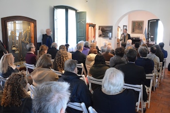 Personas sentadas en sillas blancas escuchan a un hombre que habla con un micrófono en una sala de exposición