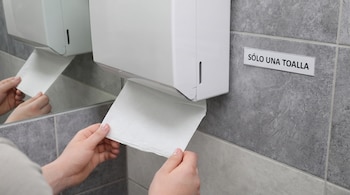 Manos tomando una toalla de papel blanca de un dispensador pegado a una pared con el cartel sólo una toalla.