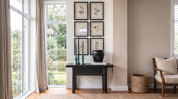 Una mesa consola negra con tres candelabros y un jarrón, seis láminas botánicas enmarcadas en la pared, un ventanal y un sillón con una cesta.