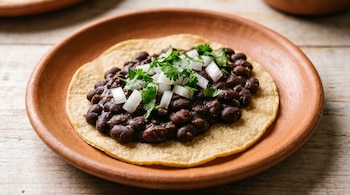 Un taco de frijol negro abierto sobre una tortilla de maíz en un plato de barro. Está cubierto con cebolla blanca picada y cilantro fresco, sobre una mesa de madera clara.