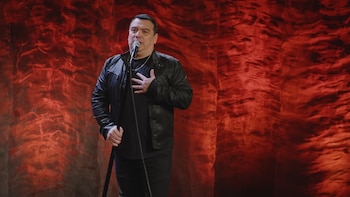 Comediante Carlos Mencia de pie en un escenario con fondo rojo, sosteniendo un micrófono con la mano izquierda y la mano derecha en el pecho. Viste chaqueta negra