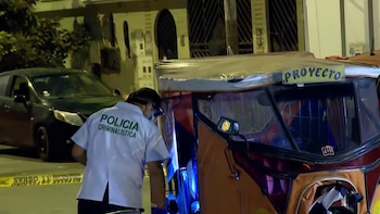 Un agente de la Policía Criminalística inspecciona una mototaxi naranja con un cartel de 'PROYECTO' en la parte superior. Cinta policial en el suelo y un auto oscuro detrás