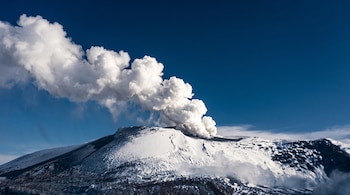 Un volcán cubierto de nieve emite una gran nube blanca de humo hacia el cielo azul.