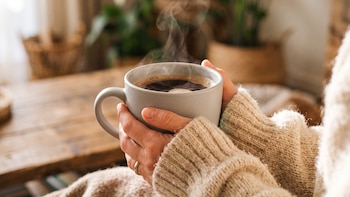 Dos manos con suéter tejido sostienen una taza gris llena de café negro humeante sobre un fondo difuminado de madera y plantas
