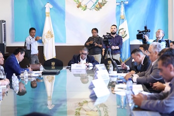 Hombres sentados en una mesa de reuniones con botellas de agua mineral, banderas de Guatemala al fondo y periodistas grabando el evento