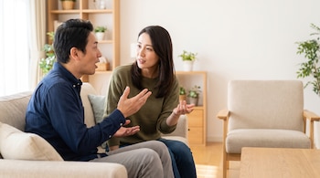 Dos adultos sentados en un sofá conversando. Un hombre gesticula con la boca abierta. Una mujer mira al hombre. Fondo con estantería de madera y plantas.