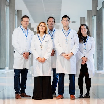 Cinco profesionales médicos, tres hombres y dos mujeres, vestidos con batas blancas y credenciales azules, posan de pie en un pasillo luminoso