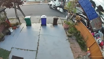 Camión de basura blanco con volquete azul volcando residuos en un jardín residencial junto a una cerca de madera. Se ven tres contenedores de basura en la entrada