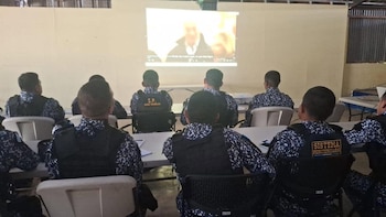 Grupo de personas con uniformes y chalecos con el texto 'Sistema Penitenciario' sentadas en un salón, observando una proyección de video