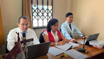 Tres personas, dos hombres y una mujer, se encuentran sentadas alrededor de una mesa de madera con tres ordenadores portátiles, documentos, un micrófono y un crucifijo