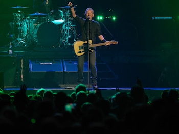 Bruce Springsteen canta y toca la guitarra en el escenario, con un brazo levantado, frente a una batería y un amplificador Marshall. El público en primer plano. Luces verdes