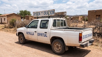 Camioneta blanca con letreros de Patrulla Espiritual y Ayuda y oración estacionada en un camino de tierra cerca de casas rurales de adobe.