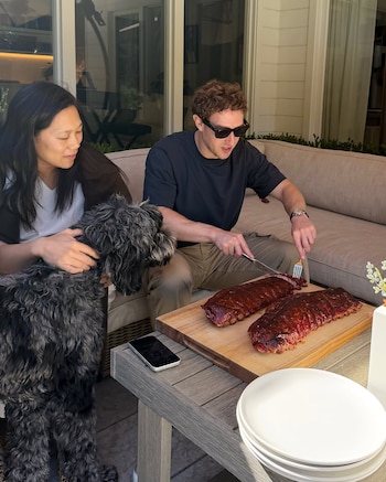 Mark Zuckerberg con gafas de sol corta costillas en una tabla junto a Priscilla Chan, un perro de pelo rizado, un teléfono y varios platos