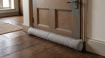 Un burlete cilíndrico de tela gris con diseño de espiga descansa sobre el suelo de madera, pegado a la base de una puerta de madera.