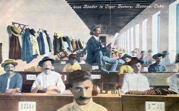 Un hombre en traje lee en alto a filas de trabajadores con bigote sentados en pupitres, elaborando puros en una fábrica cubana, con ropa colgada al fondo