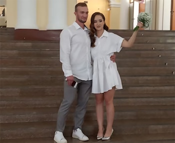 Un hombre y una mujer vestida de blanco que sostiene un ramo de flores posan juntos en la escalinata de un edificio