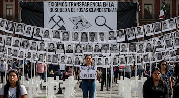 Mujer sostiene cartel 'Verdad y Justicia', rodeada de cruces blancas y fotografías. Al fondo, pancarta exige 'Transparencia en la búsqueda de fosas clandestinas'.