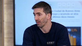 Daniel Lauretta, con barba y camiseta azul, lleva un micrófono de diadema mientras mira hacia la izquierda. Se observa una pantalla con texto al fondo