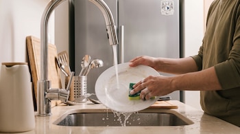 Manos de un hombre lavando un plato blanco con agua corriente y una esponja con espuma en un fregadero de acero inoxidable.