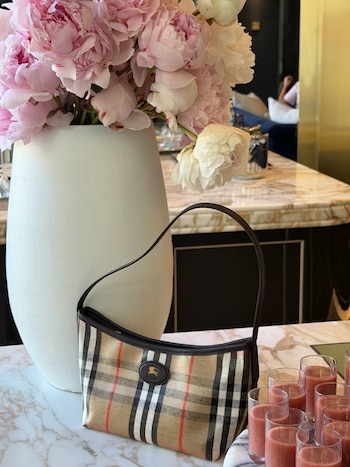 Bolso beige a cuadros sobre mesa de mármol, junto a jarrón blanco con peonías rosas y blancas. Varias copas con bebida rosada al lado