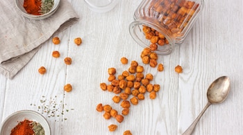 Garbanzos tostados derramados de un frasco de vidrio, cuencos con especias, una servilleta de tela y una cuchara sobre madera blanca.