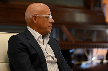 Hombre calvo de perfil con gafas transparentes, saco oscuro y camisa a rayas sentado en una silla clara con fondo desenfocado