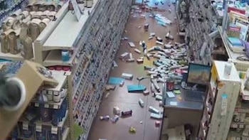 Pasillo de tienda con estantes llenos y numerosos productos desparramados en el suelo. Se observa un ordenador portátil en un mostrador