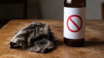 Un trapo sucio y arrugado de color oscuro y una botella de vidrio marrón con una etiqueta blanca y un círculo rojo tachado sobre mesa de madera.