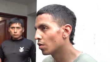 Un hombre de perfil con camiseta verde permanece junto a un marco blanco mientras otro hombre con polo negro lo observa en el fondo