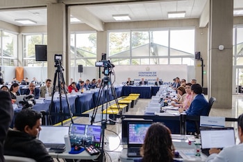 Personas sentadas en mesas dispuestas en U, con cámaras de televisión y computadoras portátiles en el área de prensa en primer plano