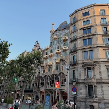 Fachada de la Casa Batlló con sus balcones y tejado escamado, rodeada de edificios adyacentes, árboles, farolas y gente caminando por la calle
