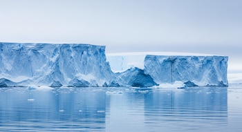 Enormes acantilados de hielo de color blanco y azul celeste se alzan sobre aguas tranquilas, con reflejos claros y pequeños fragmentos de hielo flotando.