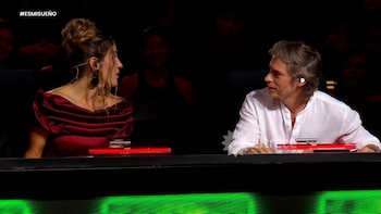 Una mujer de vestido rojo y cabello recogido mira a un hombre de camisa blanca. Ambos están sentados en un panel de evaluación con micrófonos