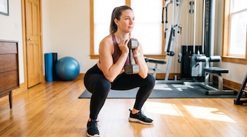 Mujer en ropa deportiva, haciendo una sentadilla. Sostiene una mancuerna. Al fondo hay aparatos de gimnasio, una pelota azul y una puerta de madera.